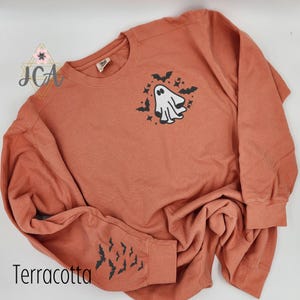 May include: Terracotta-colored sweatshirt with a crew neck and long sleeves. The front features a white ghost graphic with black bats and stars. The left sleeve has a bat graphic. The word "Terracotta" is printed at the bottom.