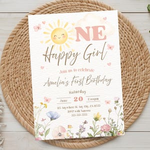 May include: A pastel-colored birthday invitation with a smiling sun, butterflies, and floral accents. The text reads "ONE Happy Girl" and invites guests to Amelia's first birthday on June 20th at 2:00 PM. The invitation is set on a woven placemat.