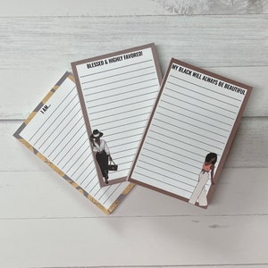 May include: Three lined notepads with inspirational quotes and illustrations of stylish women. The notepads have brown borders and gold and gray patterned accents. The text includes "I AM...", "BLESSED & HIGHLY FAVORED!", and "MY BLACK WILL ALWAYS BE BEAUTIFUL".