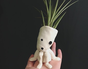 May include: A whimsical, handmade white ceramic air plant holder in the shape of a seated figure with a surprised expression. The holder contains a green air plant with long, slender leaves. The figure has two black dot eyes and a surprised open mouth.