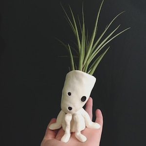 Puede incluir: Un soporte para plantas aéreas de cerámica blanca hecho a mano con forma de figura sentada con expresión de sorpresa. El soporte contiene una planta aérea verde con hojas largas y delgadas. La figura tiene dos ojos de puntos negros y una boca abierta.