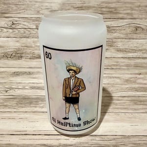 May include: Frosted glass can-shaped tumbler with a colorful graphic. The graphic features a cartoon figure in a brown jacket and shorts, holding a football. The number "60" is in the upper left corner, and the text "El Halftime Show" is below the figure.