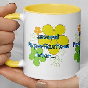 Op de afbeelding: Witte keramische mok met een gele rand. De mok heeft een kleurrijk bloemenmotief en de tekst "several hyperfixations later..." in blauw.