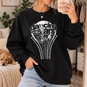 May include: A black crewneck sweatshirt featuring a white graphic print of a disco ball with the text "dancing with the stars" centered on the chest. The sweatshirt has long sleeves with ribbed cuffs and a ribbed hem, paired with light blue denim jeans.