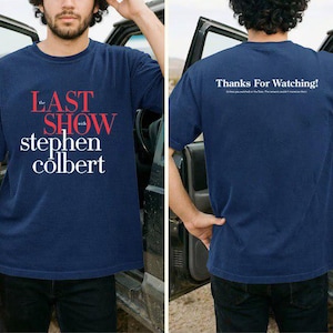 May include: Navy blue t-shirt featuring the text "The Last Show with Stephen Colbert" in red and white. The back of the shirt displays "Thanks For Watching!" in white text. The shirt is a short-sleeved crew neck.