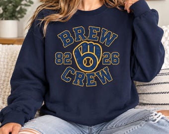 May include: A navy blue crewneck sweatshirt featuring a distressed yellow and blue graphic print that reads BREW CREW with the numbers 82 and 26 on either side of a central baseball glove icon containing a baseball.