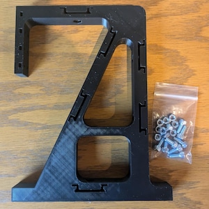 May include: A black, L-shaped bracket with a triangular support structure and multiple rectangular cutouts along its edges. The bracket features a series of small square holes for mounting. A small clear plastic bag contains silver metal bolts and nuts.