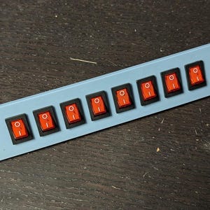 May include: A pale blue panel with eight black rectangular switches, each with a red toggle. The switches are arranged in a row. The panel has two small holes on the left and right sides.