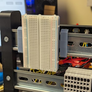 May include: A close-up of a breadboard circuit board with rows of holes for electronic components. The board is white with blue and red lines and is mounted on a black metal frame. Wires are visible.