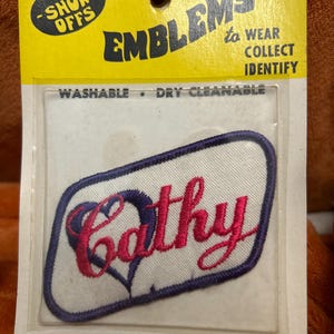 May include: Embroidered "Cathy" name patch with a heart design in pink and purple. The patch is on a white background with a purple border, packaged in a yellow and clear plastic card. The packaging reads "EMBLEMS to WEAR COLLECT IDENTIFY".