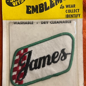 May include: Embroidered name patch with the name "James" in black script. The patch has a white background, a red and black checkered design, and a green border. The packaging reads "The Show-Offs" and "Emblems to Wear, Collect, Identify."