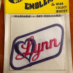 May include: Embroidered patch with the name "Lynn" in pink script, framed by a purple heart design and border, on a white background. The patch is packaged in a clear plastic sleeve with a yellow backing that reads "EMBLEMS to WEAR COLLECT IDENTIFY".
