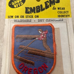 May include: Embroidered Oregon patch with a blue background, orange border, and a lumberjack design. The patch is packaged with a yellow backing that reads "The Show-Offs" and "Emblems to Wear, Collect, Identify."