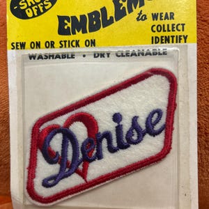 May include: Embroidered patch with the name "Denise" in blue script, framed by a red border on a white background. The patch is packaged on a yellow card with the text "EMBLEMS to WEAR COLLECT IDENTIFY". The patch is washable and dry cleanable.