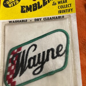 May include: Embroidered "Wayne" patch with a green border and a red and white checkered design on the left side. The patch is on a clear plastic backing, with a yellow cardboard header that reads "EMBLEMS to WEAR COLLECT IDENTIFY".