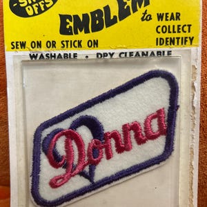 May include: Embroidered patch with the name "Donna" in pink script, outlined in purple, on a white background. The patch is in a purple rectangular shape. The packaging reads "EMBLEMS to WEAR COLLECT IDENTIFY".