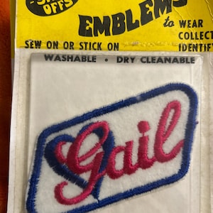 May include: Embroidered patch with the name "Gail" in pink script, outlined in blue, on a white background. The patch is rectangular with a blue border. The packaging reads "EMBLEMS to WEAR COLLECT IDENTIFY".