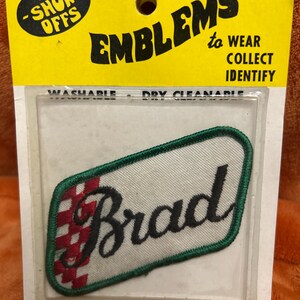 May include: Embroidered name patch with the name "Brad" in black script on a white background. The patch has a green border and a red and black checkered design on the left side. The packaging reads "EMBLEMS to WEAR COLLECT IDENTIFY".