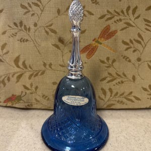 May include: A blue glass bell-shaped cologne bottle with a silver-colored top and stopper. The bottle has a diamond pattern and a label that reads "Moonwind Cologne". The bottle is 3.75 fl oz.