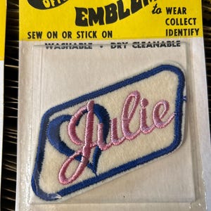 May include: Embroidered patch with the name "Julie" in pink script, outlined in blue, on a white background. The patch is in a clear plastic package with a yellow header that reads "EMBLEMS to WEAR COLLECT IDENTIFY".