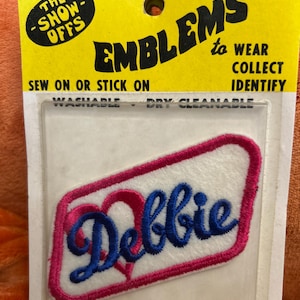 May include: Embroidered "Debbie" patch with a pink heart design and blue lettering, on a white background. The patch is packaged on a yellow card with the text "The Show-Offs" and "Emblems to Wear, Collect, Identify."