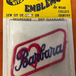 May include: Embroidered "Barbara" patch with a pink heart design and a white background, encased in a clear plastic package. The package has a yellow backing with the text "The Show-Offs" and "Emblems to Wear, Collect, Identify".