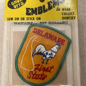 May include: Embroidered Delaware state emblem patch. The patch features a rooster, the state outline, and the words "Delaware" and "First State". The patch is on a yellow backing with green trim and is packaged in a clear plastic sleeve.