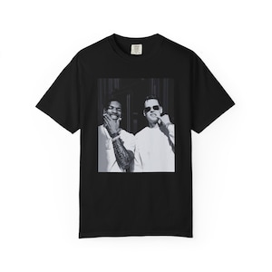 May include: Black t-shirt featuring a black and white image of two people. One person is wearing sunglasses and the other is smiling. The t-shirt is made of a soft material.