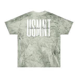 May include: A light green tie-dye t-shirt featuring the text "USMNT" in large white capital letters, with "Christian Pulisic" in smaller white script below. The shirt has a crew neck and short sleeves.