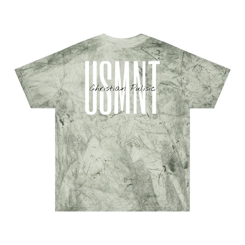 May include: A light green tie-dye t-shirt featuring the text "USMNT" in large white capital letters, with "Christian Pulisic" in smaller white script below. The shirt has a crew neck and short sleeves.