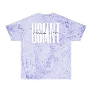 May include: A light purple tie-dye t-shirt featuring the text "USMNT" stacked vertically and "Christian Pulisic" below it in white. The shirt has a crew neck and short sleeves.