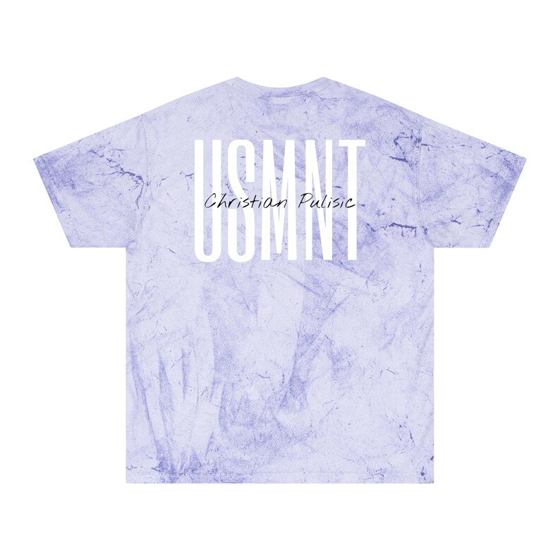 May include: A light purple tie-dye t-shirt featuring the text "USMNT" stacked vertically and "Christian Pulisic" below it in white. The shirt has a crew neck and short sleeves.