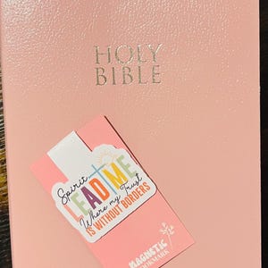 May include: A pink Holy Bible with embossed gold lettering and a pink magnetic bookmark. The bookmark has the words "Lead Me" and "Spirit" printed on it, along with the words "Where my Trust is Without Borders."