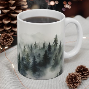 May include: White ceramic mug featuring a forest scene with tall evergreen trees and a misty mountain backdrop. The mug is filled with a dark liquid and has a curved handle. The mug is surrounded by pine cones and a lit candle.