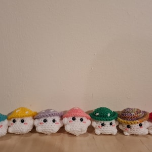 May include: A row of eight crocheted mushroom plushies in various colours, including teal, yellow, purple, pink, green, multi-coloured, and red.