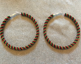 May include: A pair of hoop earrings with a beaded design. The earrings feature a pattern of red, green, and black beads. The hoops are made of silver-colored metal and are displayed on a gray surface.