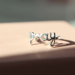 May include: A silver ring spelling out the word "pray" in a cursive style. The ring sits on a pale pink surface, with a shadow to the right. The ring is a piece of jewellery.