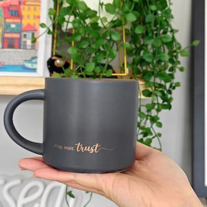 May include: A matte charcoal gray ceramic mug with a rounded handle. The front of the mug features the text "Pray, wait. trust" printed in a metallic copper-colored script font. The mug has a smooth, solid-colored finish and a cylindrical shape.