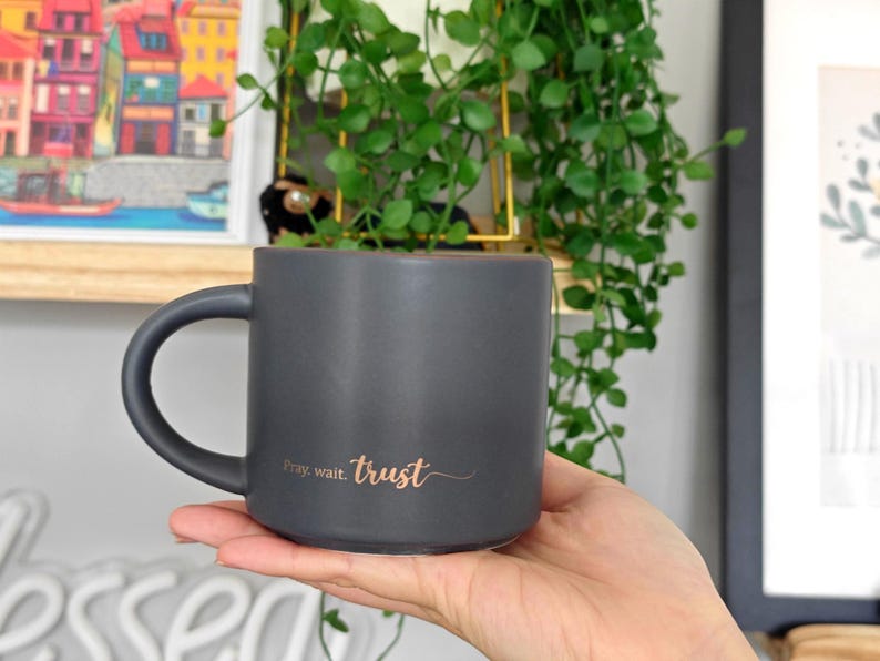 May include: A matte charcoal gray ceramic mug with a rounded handle. The front of the mug features the text "Pray, wait. trust" printed in a metallic copper-colored script font. The mug has a smooth, solid-colored finish and a cylindrical shape.