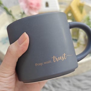 May include: A matte charcoal gray ceramic mug with a rounded handle and a flat base. The side of the mug features the text "Pray. wait. trust" printed in a metallic gold script font. The mug has a smooth, solid-colored exterior finish.