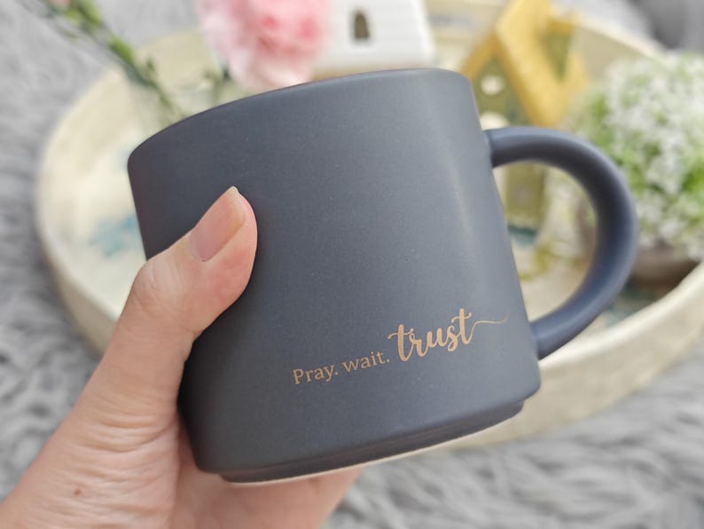 May include: A matte charcoal gray ceramic mug with a rounded handle and a flat base. The side of the mug features the text "Pray. wait. trust" printed in a metallic gold script font. The mug has a smooth, solid-colored exterior finish.
