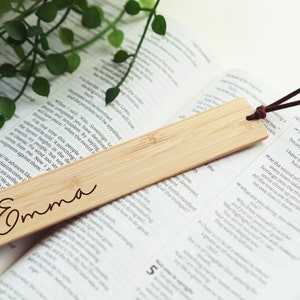 May include: A natural bamboo bookmark with the name "Emma" engraved on it. The bookmark has a brown leather string attached to it.