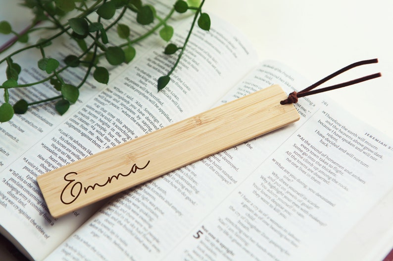 May include: A natural bamboo bookmark with the name "Emma" engraved on it. The bookmark has a brown leather string attached to it.