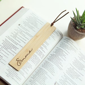 May include: A wooden bookmark with the name "Emma" engraved on it. The bookmark is light brown and has a brown leather tassel attached to it. The bookmark is lying on an open book.