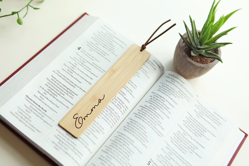 May include: A wooden bookmark with the name "Emma" engraved on it. The bookmark is light brown and has a brown leather tassel attached to it. The bookmark is lying on an open book.