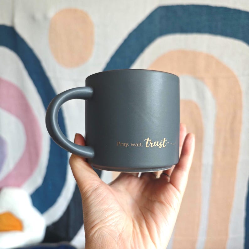 May include: A matte charcoal gray ceramic mug with a rounded handle. The side of the mug features the gold-colored text "Pray. wait. trust" written in a cursive script font. The mug has a smooth, solid-colored finish and a cylindrical shape.