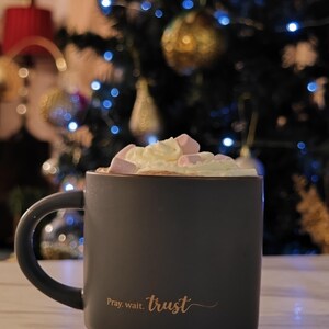 May include: A matte dark gray ceramic mug filled with whipped cream and pink marshmallows. The mug features the gold-colored text "Pray. wait. trust" printed on the front. The handle is curved and positioned on the left side of the mug.