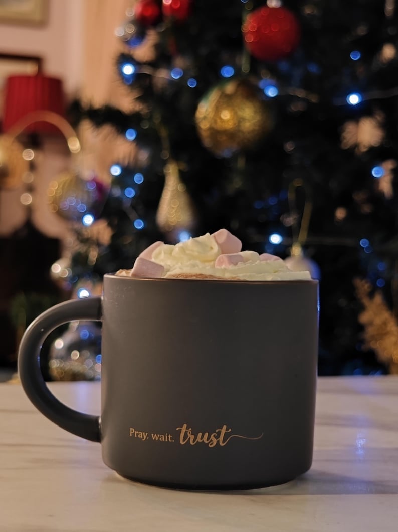 May include: A matte dark gray ceramic mug filled with whipped cream and pink marshmallows. The mug features the gold-colored text "Pray. wait. trust" printed on the front. The handle is curved and positioned on the left side of the mug.