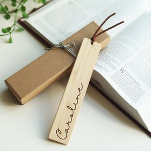 May include: A personalized wooden bookmark with the name "Caroline" engraved on it. The bookmark is brown and has a string attached to it. It is sitting on top of a closed book.