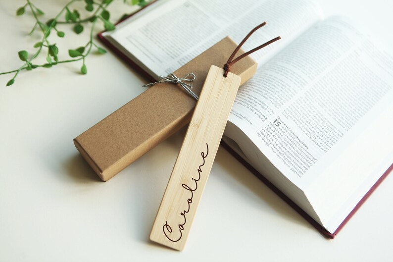 May include: A personalized wooden bookmark with the name "Caroline" engraved on it. The bookmark is brown and has a string attached to it. It is sitting on top of a closed book.